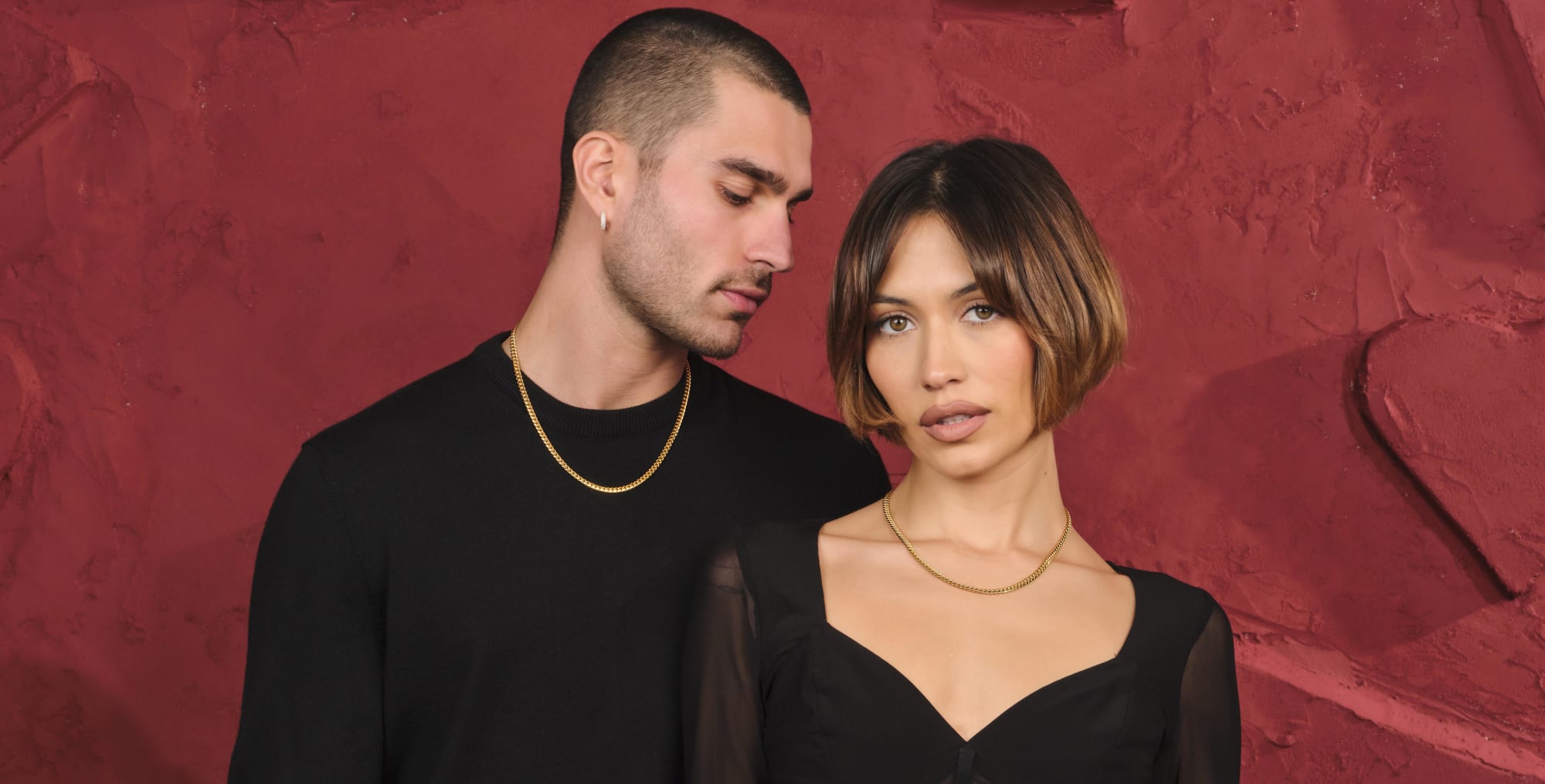 A male and female model standing together wearing the gold JAXXON His and Hers Cuban 5mm Chain Set against a red backdrop.