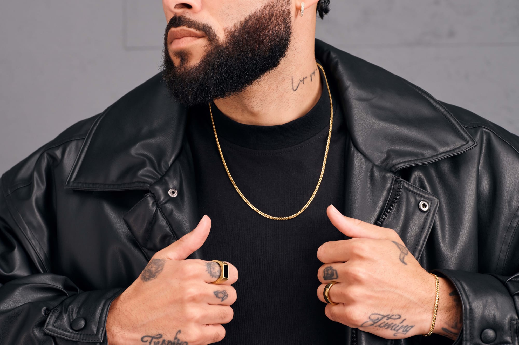 A male model clutching the lapels of his black leather jacket, wearing the JAXXON gold Cuban Link Chain and JAXXON rings.