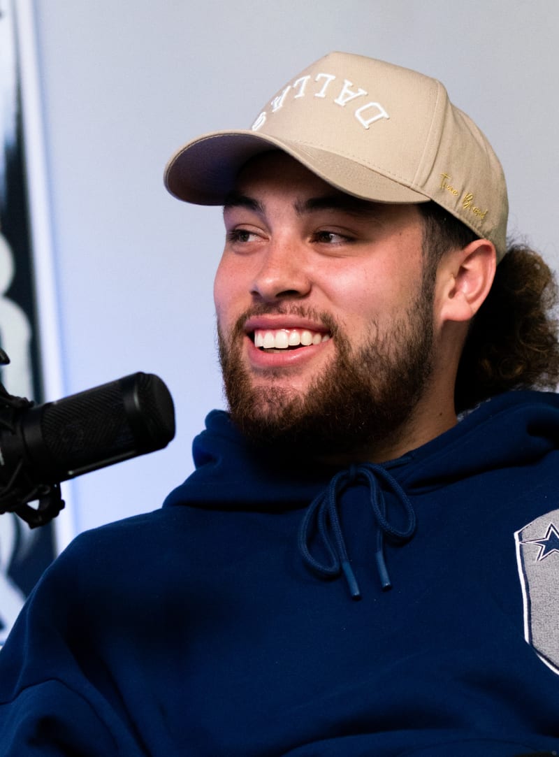 Simi Fehoko sitting in the JAXXON Podcast Studio smiling in front of a microphone, in a black hoodie and beige baseball cap.