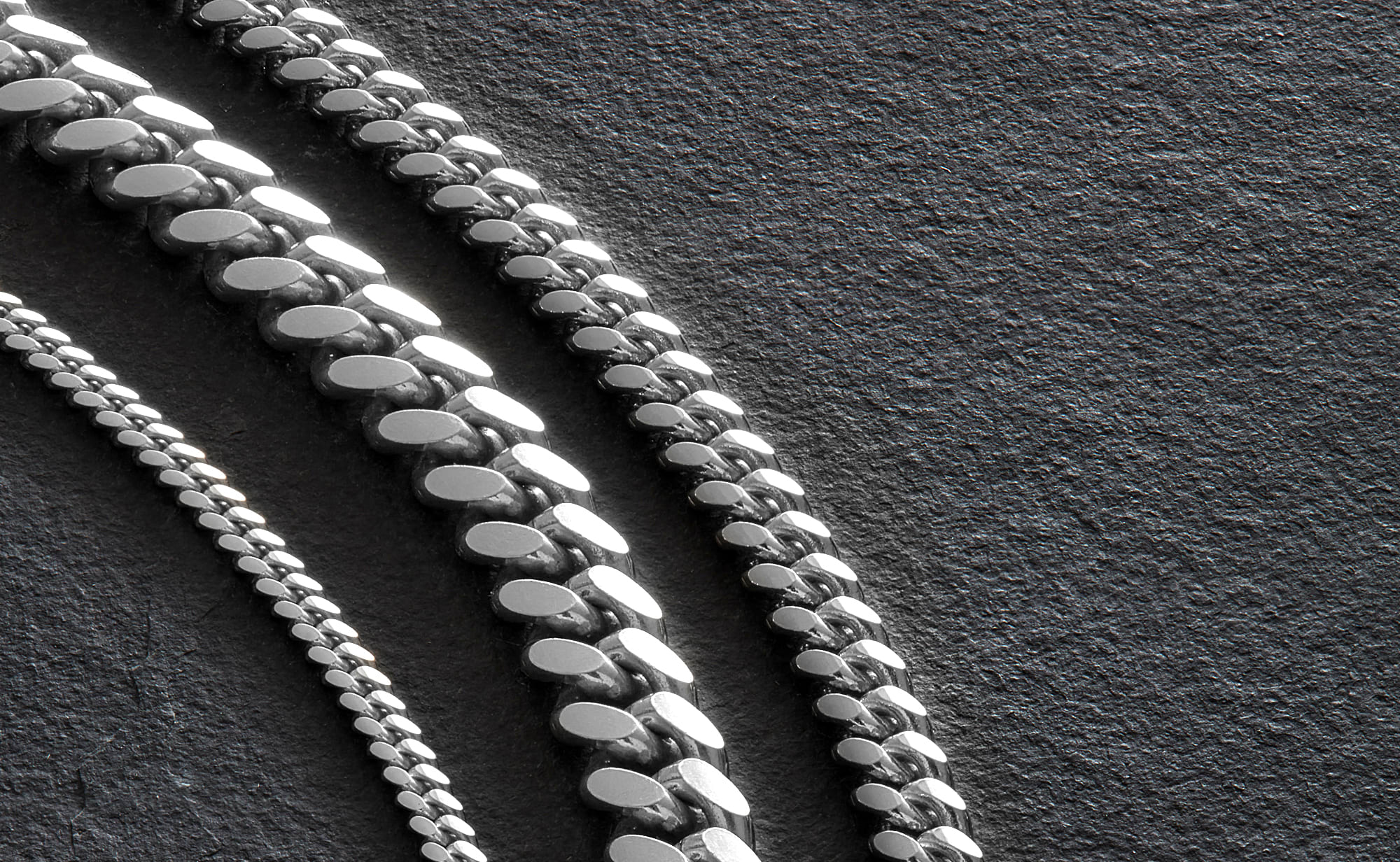 A close up image of the JAXXON 3mm, 7mm and 5mm Silver Cuban Link Chains lying on a black, textured stone surface.