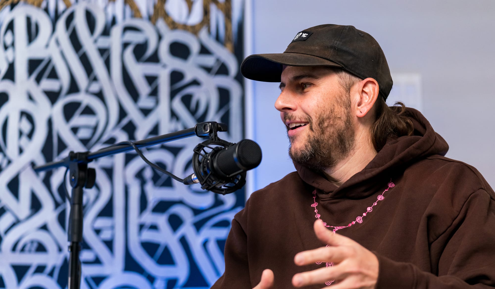 M Shadows on a white couch in the JAXXON Podcast Studio, wearing a black hoodie and black ball cap, talking into the microphone.