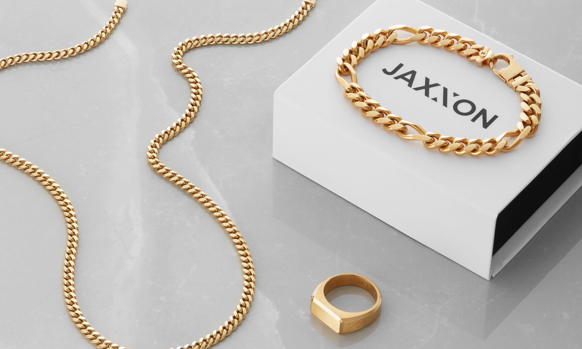 JAXXON's Gold Cuban Link Chain, Figaro Bracelet, and Signet Ring laying on a grey marble surface with a JAXXON gift box.