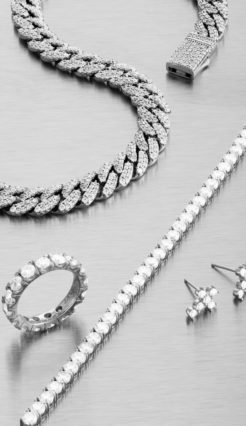 A close-up view of several silver JAXXON Iced Out jewelry pieces laid flat, including the Tennis Chain and Iced Out Cuban Necklace.