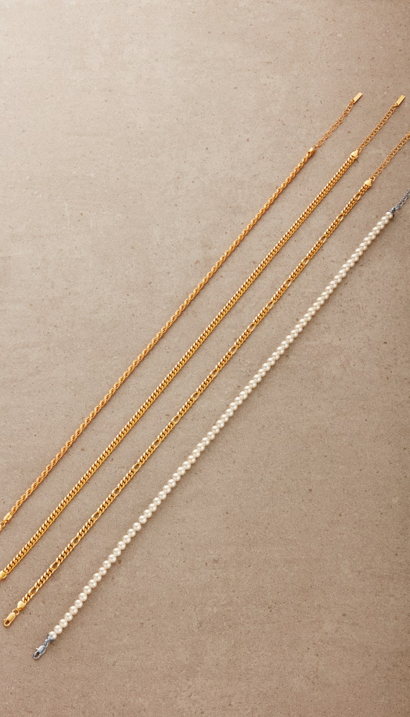 An overhead view of JAXXON's Rope, Cuban, Figaro chains and Pearl Choker laid in a straight line, side by side.