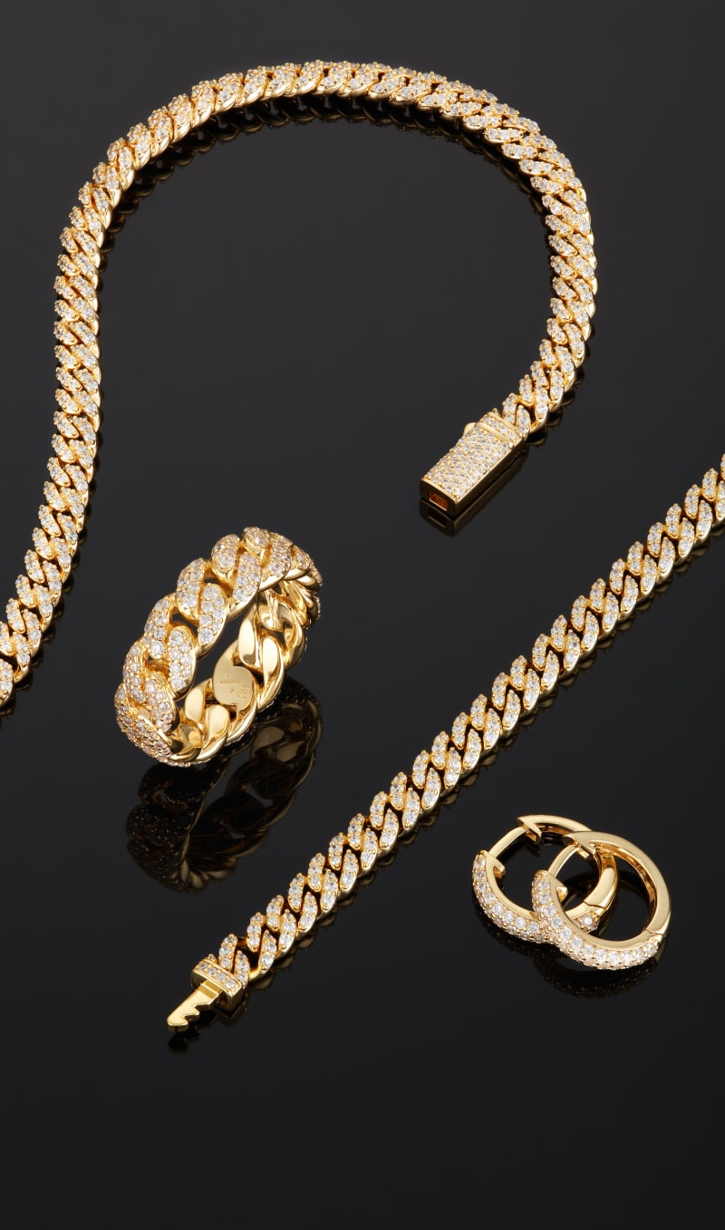 View of the gold Iced Out Cuban Essentials Set, Iced Out Cuban Link Ring, and Iced Out Hoop Earrings on a reflective, black surface.