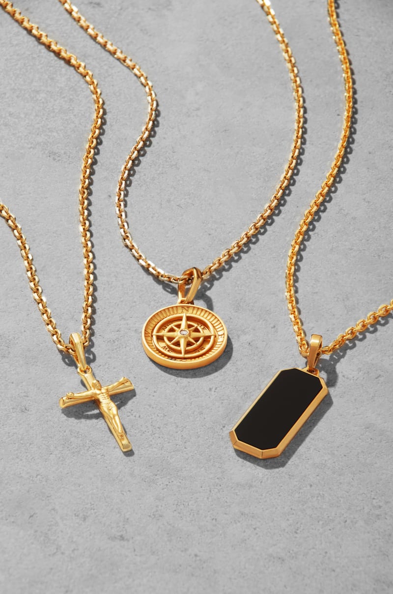 JAXXON's Gold Crucifix, Compass, and Onyx Beverly Pendants on gold cable chains laid out together on a flat grey background.
