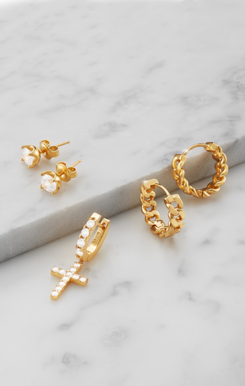 A view of JAXXON's Gold Classic Studs, Cuban Link Hoops, and a Hanging Studded Cross earring on a marble surface.