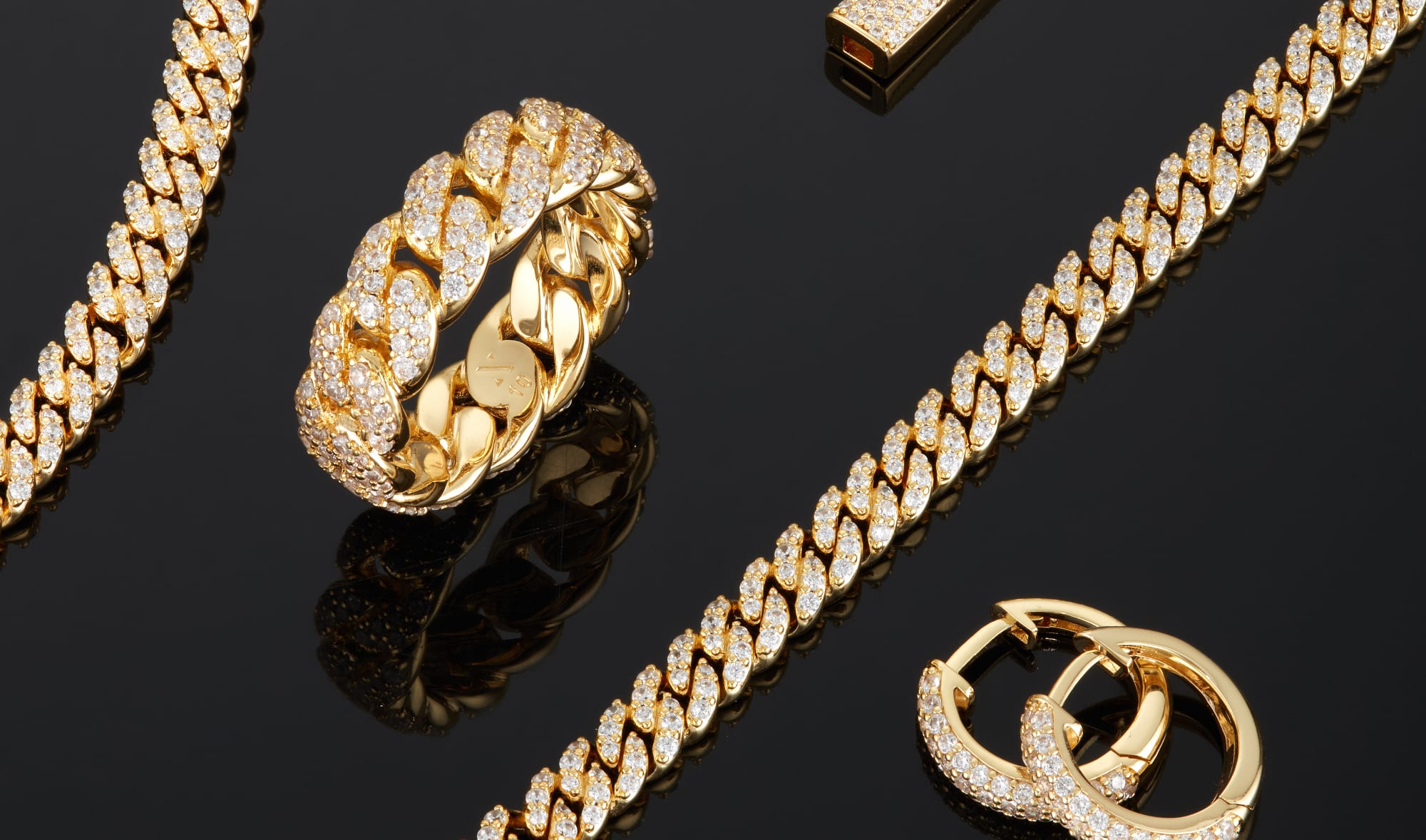 View of the gold Iced Out Cuban Essentials Set, Iced Out Cuban Link Ring, and Iced Out Hoop Earrings on a reflective, black surface.