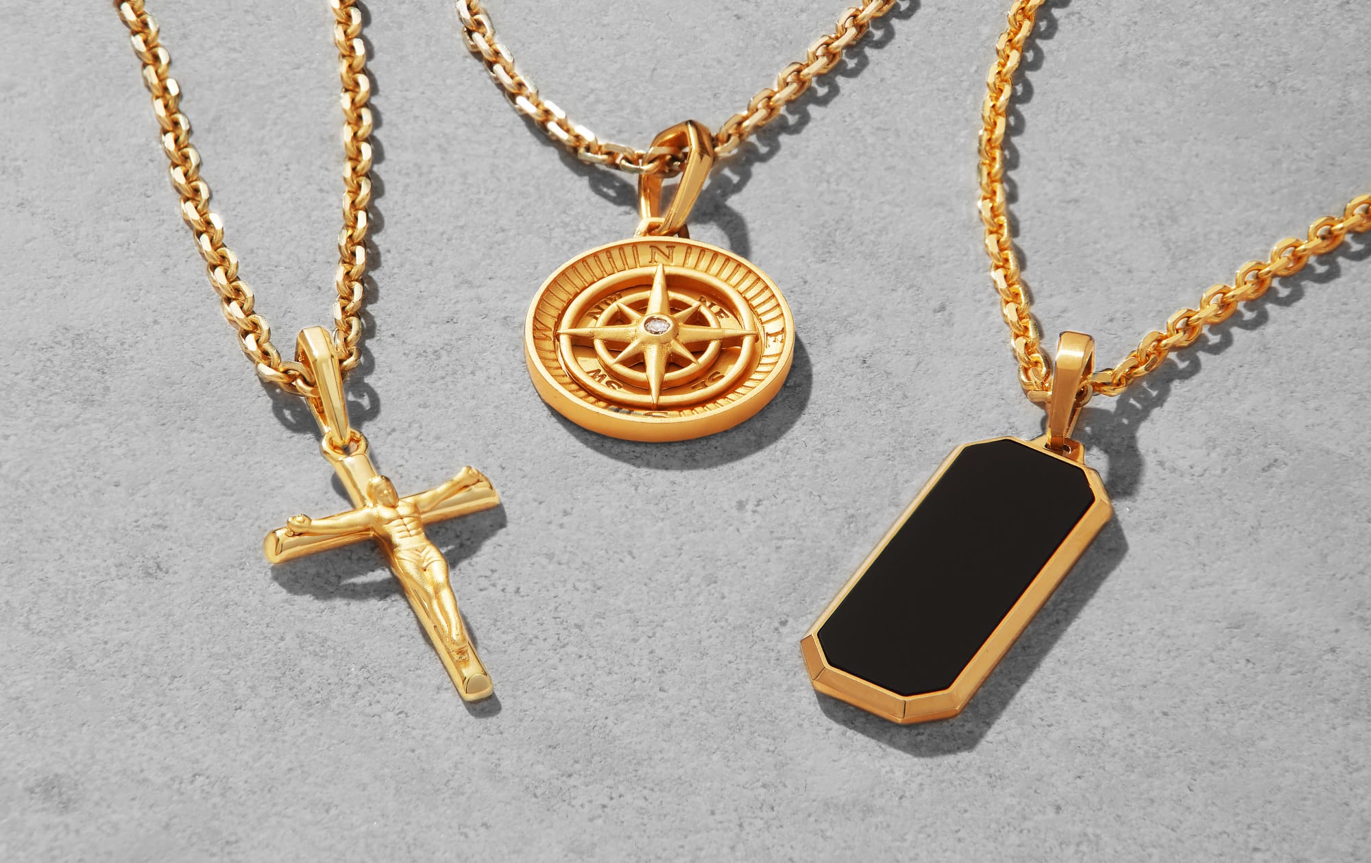 JAXXON's Gold Crucifix, Compass, and Onyx Beverly Pendants on gold cable chains laid out together on a flat grey background.
