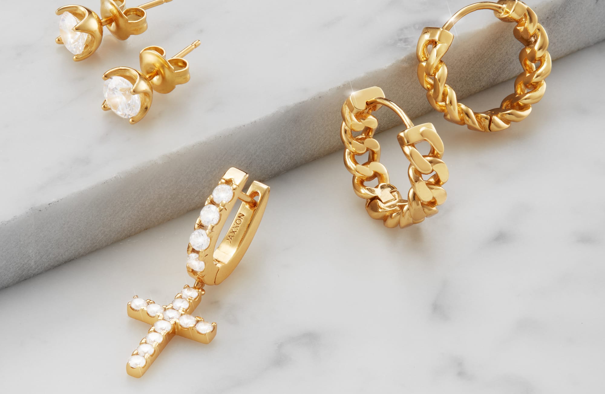 A view of JAXXON's Gold Classic Studs, Cuban Link Hoops, and a Hanging Studded Cross earring on a marble surface.