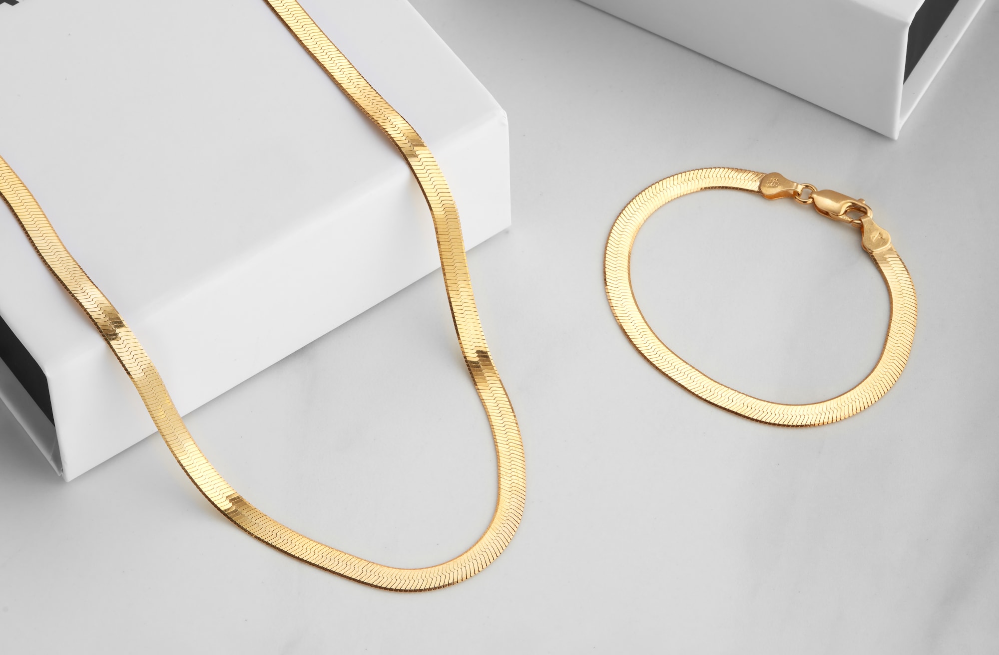 JAXXON's gold 5mm Herringbone chain descending from a white JAXXON box, with the matching bracelet by it's side, on a white marbled surface.