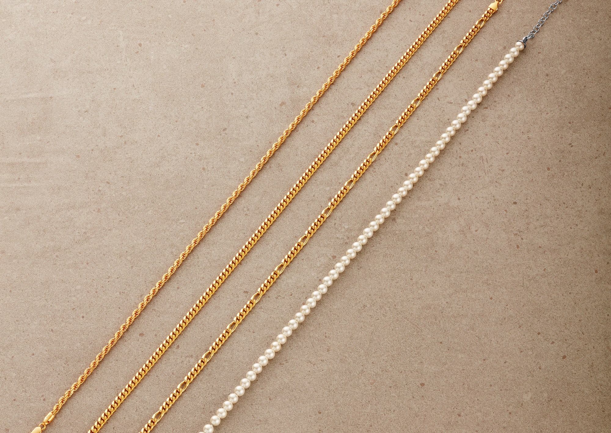An overhead view of JAXXON's Rope, Cuban, Figaro chains and Pearl Choker laid in a straight line, side by side.
