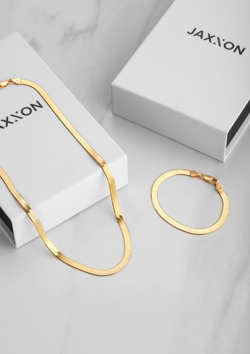 JAXXON's gold 5mm Herringbone chain descending from a white JAXXON box, with the matching bracelet by it's side, on a white marbled surface.