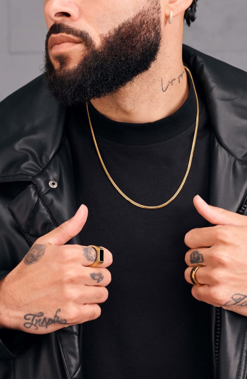 A male model clutching the lapels of his black leather jacket, wearing the JAXXON gold Cuban Link Chain and JAXXON rings.