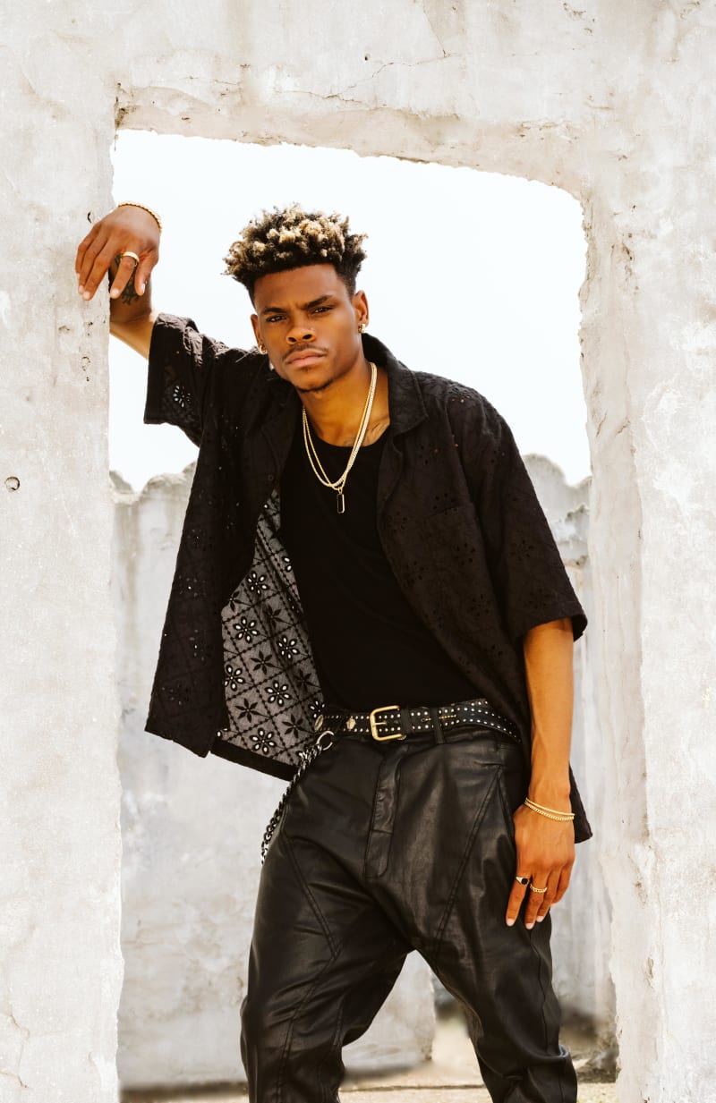 Robert Neal standing in a carved-out stone doorway, wearing all black clothes, styled with multiple JAXXON gold jewelry pieces.