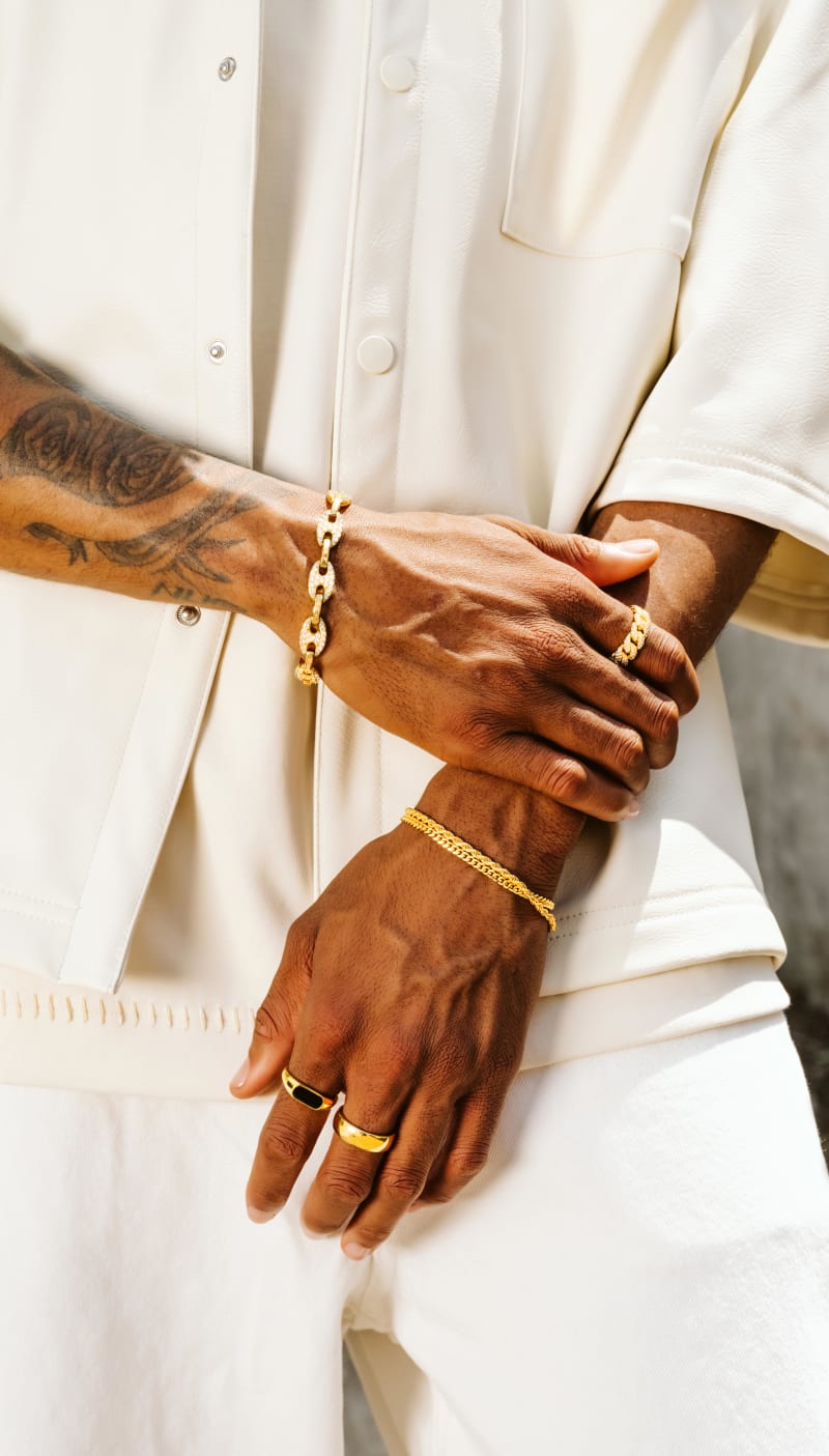 A close-up view of a male model clutching his wrist, wearing gold JAXXON jewelry pieces including the Iced Out Mariner Bracelet.