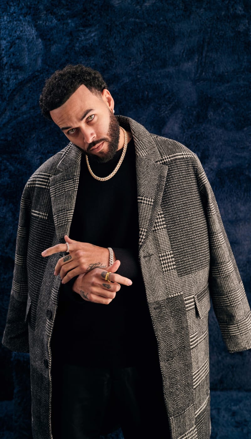 A male model wearing the JAXXON gold Iced Out Cuban Set, styled with a grey, mixed plaid peacoat draped over his shoulders.