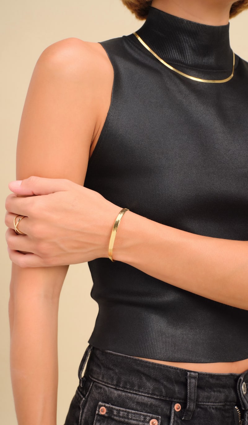 A zoomed in view of a female model, clutching her arm, wearing a black sleeveless mock neck top and the JAXXON Gold Herringbone Set.