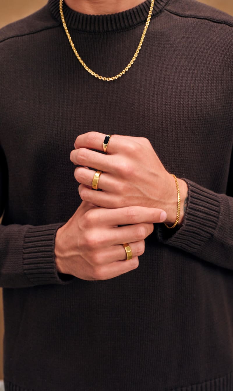 A zoomed in view of Jordan Adessi's hands, wearing a black sweater, and JAXXON's Gold Stadium Ring, Wilshire Ring, and Twelve Ring.