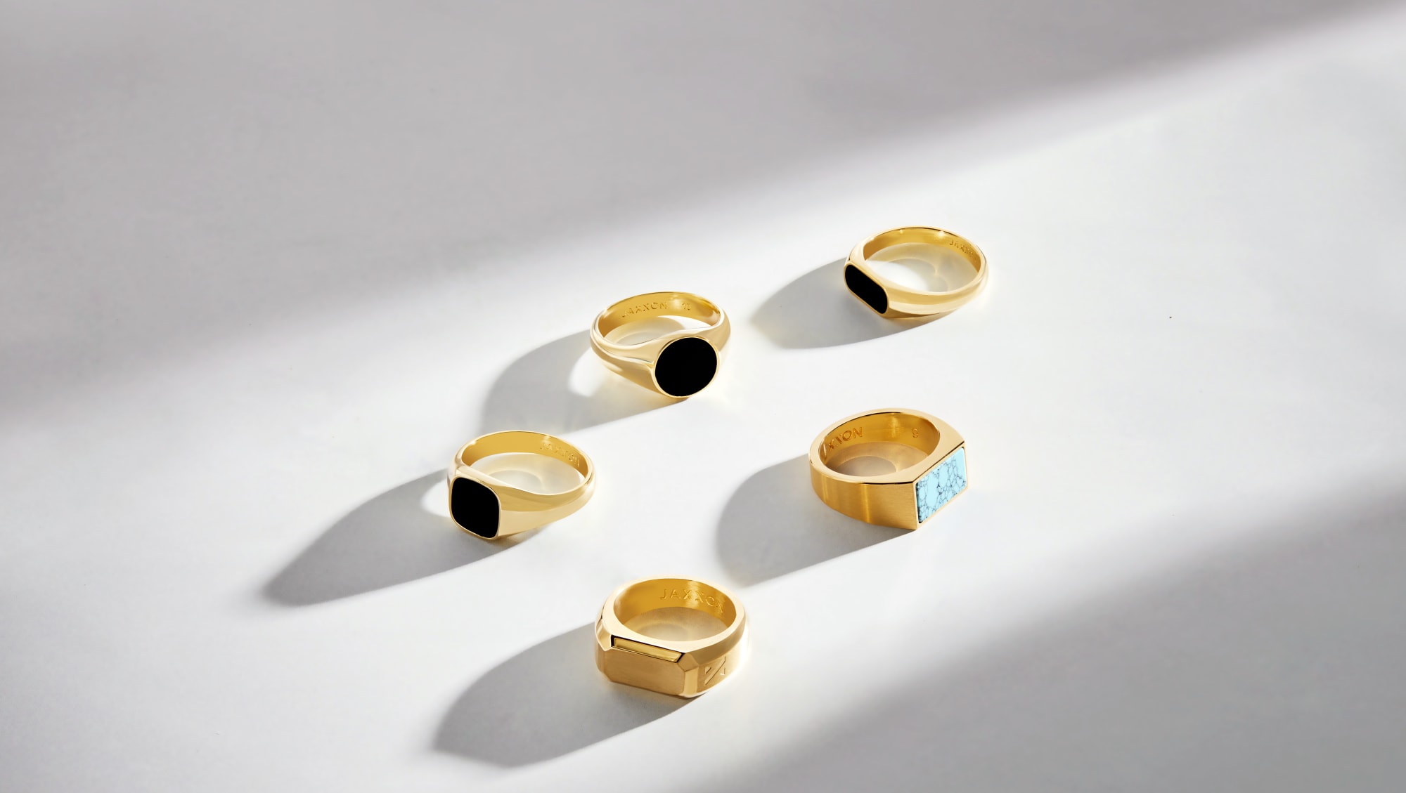 A view of five JAXXON Signet Rings on a white surface in the center of a beam of sunlight. including turquoise an onyx inlay stones.