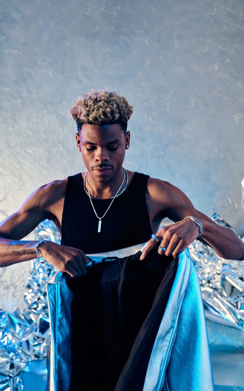 Robert Neal holding a blue metallic leather jacket in a black tank top wearing the JAXXON Silver Classic Stud Earrings, multiple JAXXON silver chains, silver rings, and silver bracelets against a metallic silver backdrop.