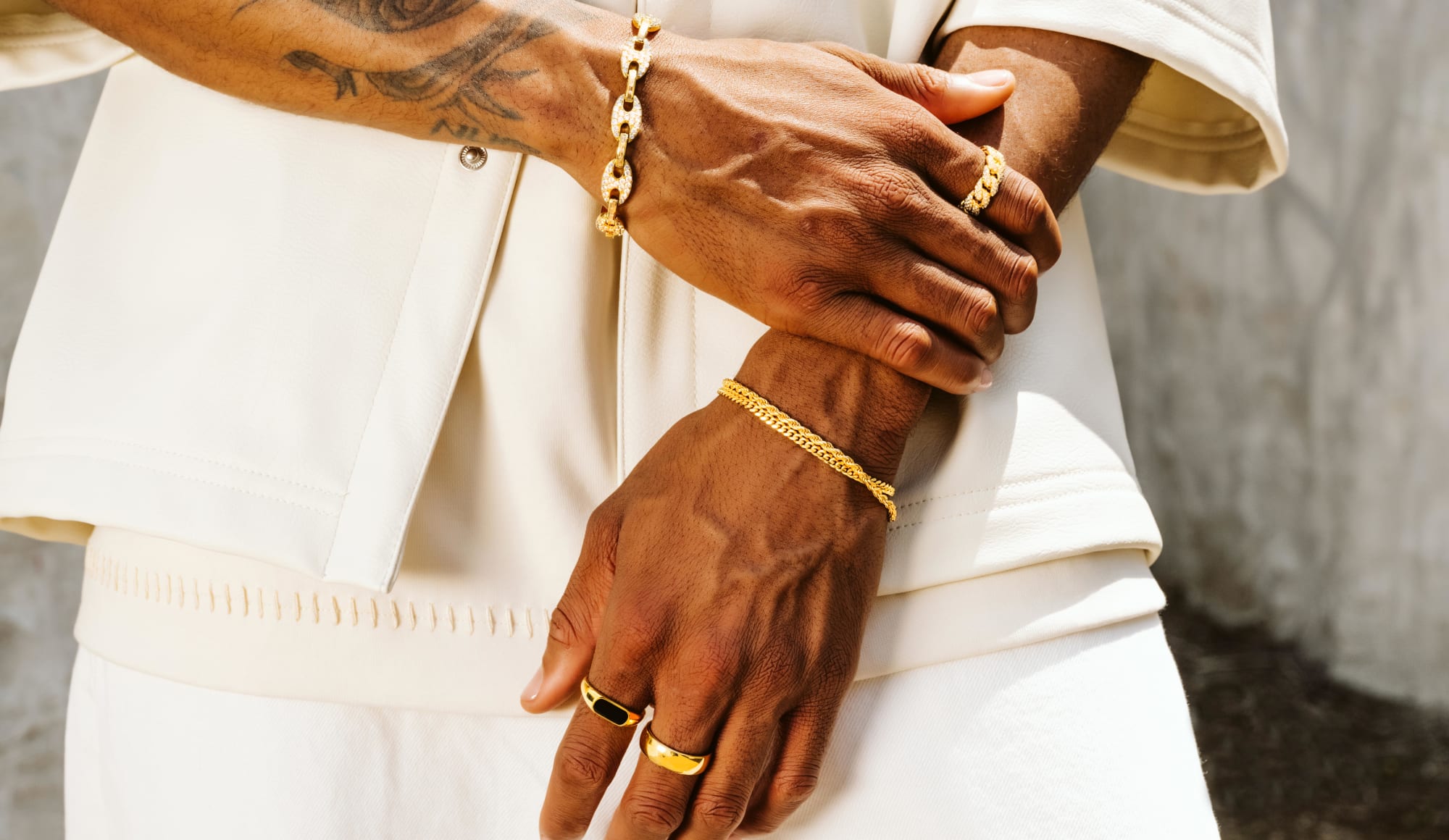 A close-up view of a male model clutching his wrist, wearing gold JAXXON jewelry pieces including the Iced Out Mariner Bracelet.
