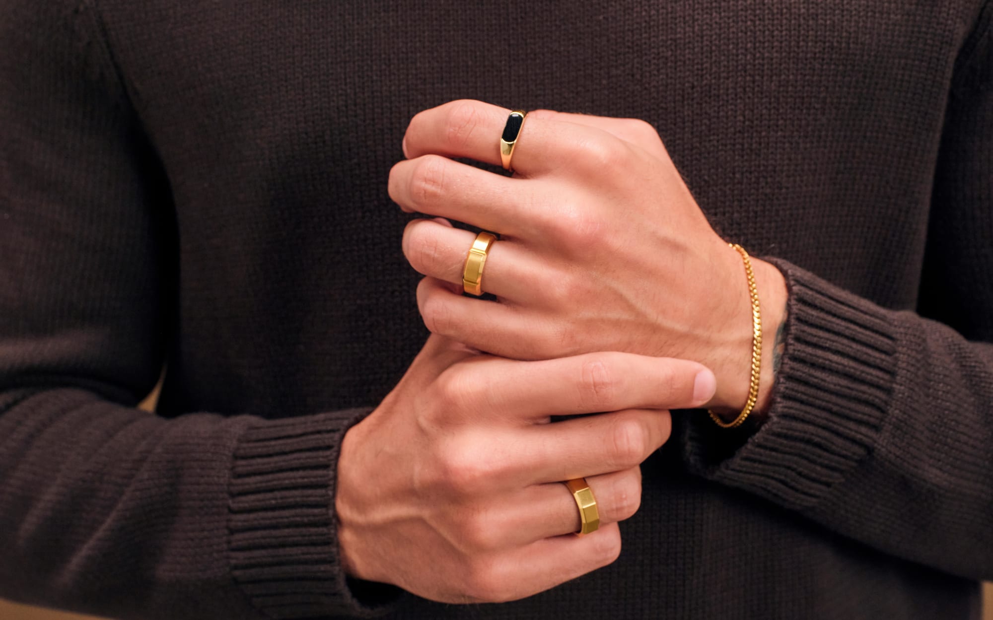 A zoomed in view of Jordan Adessi's hands, wearing a black sweater, and JAXXON's Gold Stadium Ring, Wilshire Ring, and Twelve Ring.