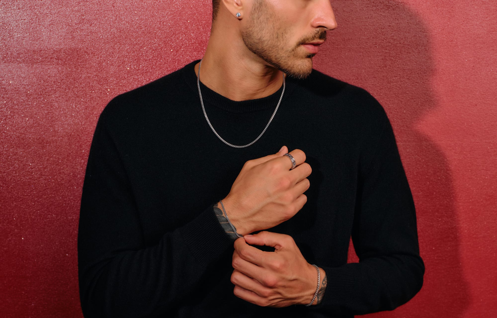 A male model sitting on a stool, adjusting his bracelet wearing all black clothing styled with the JAXXON Silver Cuban Link Bracelet.