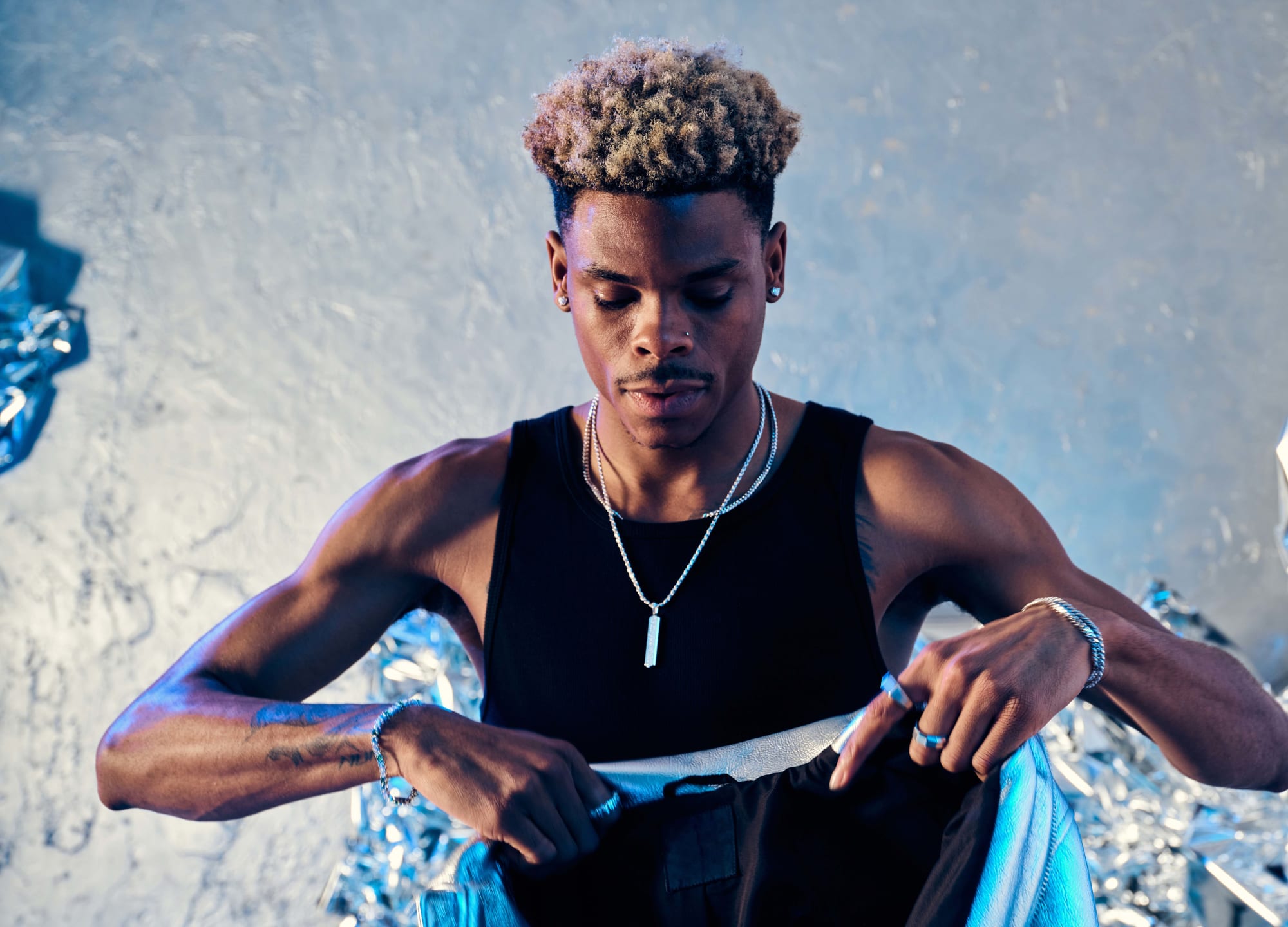 Robert Neal holding a blue metallic leather jacket in a black tank top wearing the JAXXON Silver Classic Stud Earrings, multiple JAXXON silver chains, silver rings, and silver bracelets against a metallic silver backdrop.